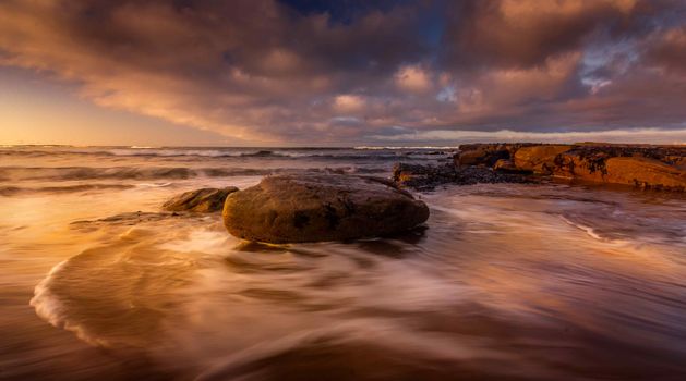 Golden sunset reflecting on ocean waves with large rocks, dramatic clouds overhead.