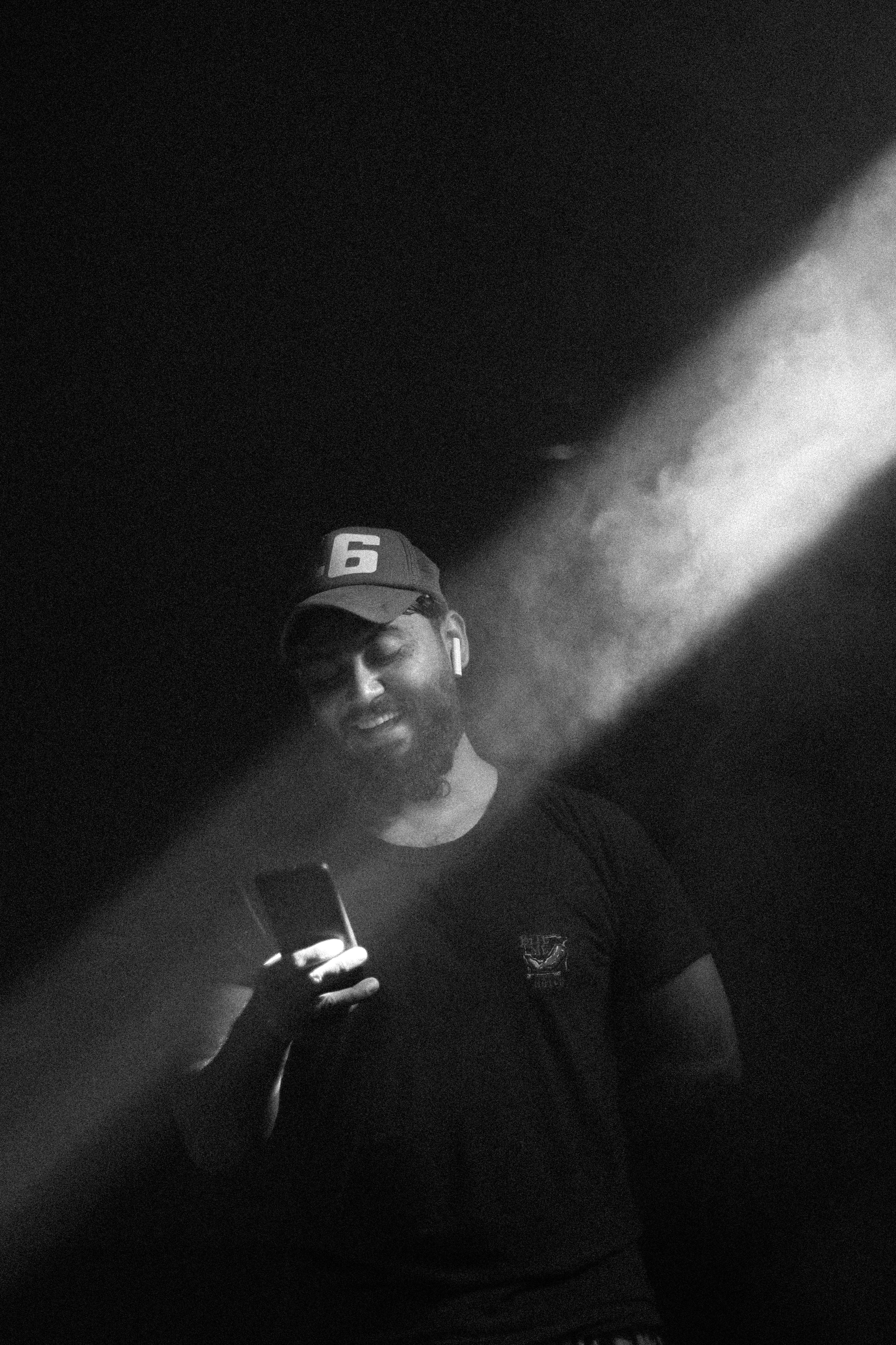 A man smiles while using a smartphone in a dark, smoke-filled room with a spotlight on him.