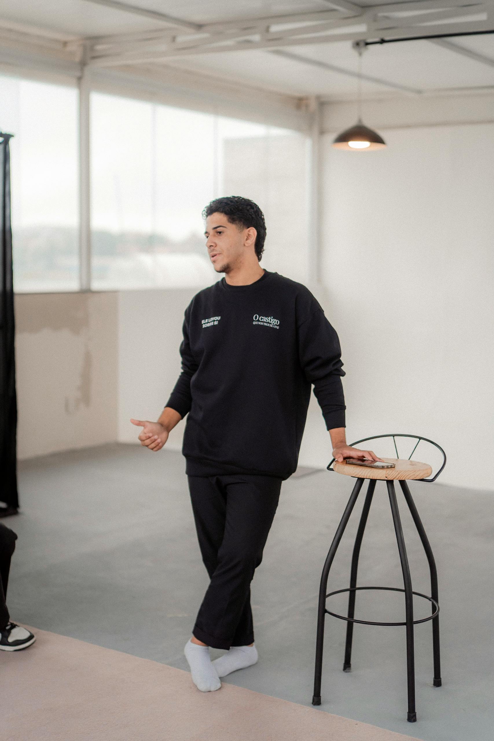 A man casually leaning on a modern stool in a bright indoor setting, conveying a relaxed atmosphere.