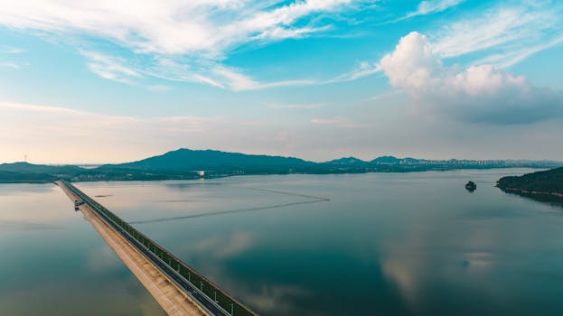 A tranquil aerial landscape of a bridge crossing a serene lake with mountains in the background under a vibrant sky.