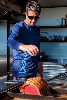 Chef in sunglasses seasoning a cooked roast in a brightly lit modern kitchen.