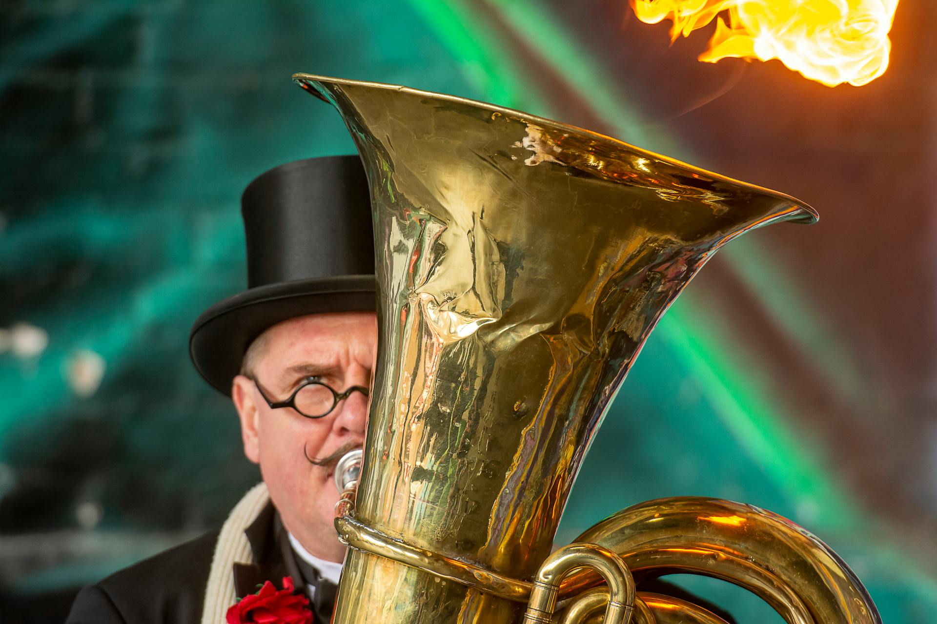 A musician with a top hat and mustache plays a tuba emitting flames, captured in a vibrant performance setting.