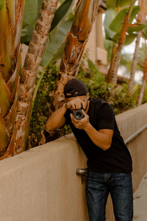 Free A man photographing near palm trees, embodying outdoor creativity and focus. Stock Photo