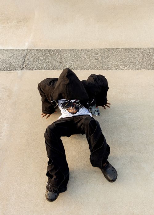 Free A teenager in a black hooded outfit sits on concrete outdoors, exuding urban style and attitude. Stock Photo