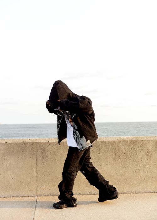 Free A person in urban attire walks along a seaside promenade, hood pulled up against the breeze. Stock Photo