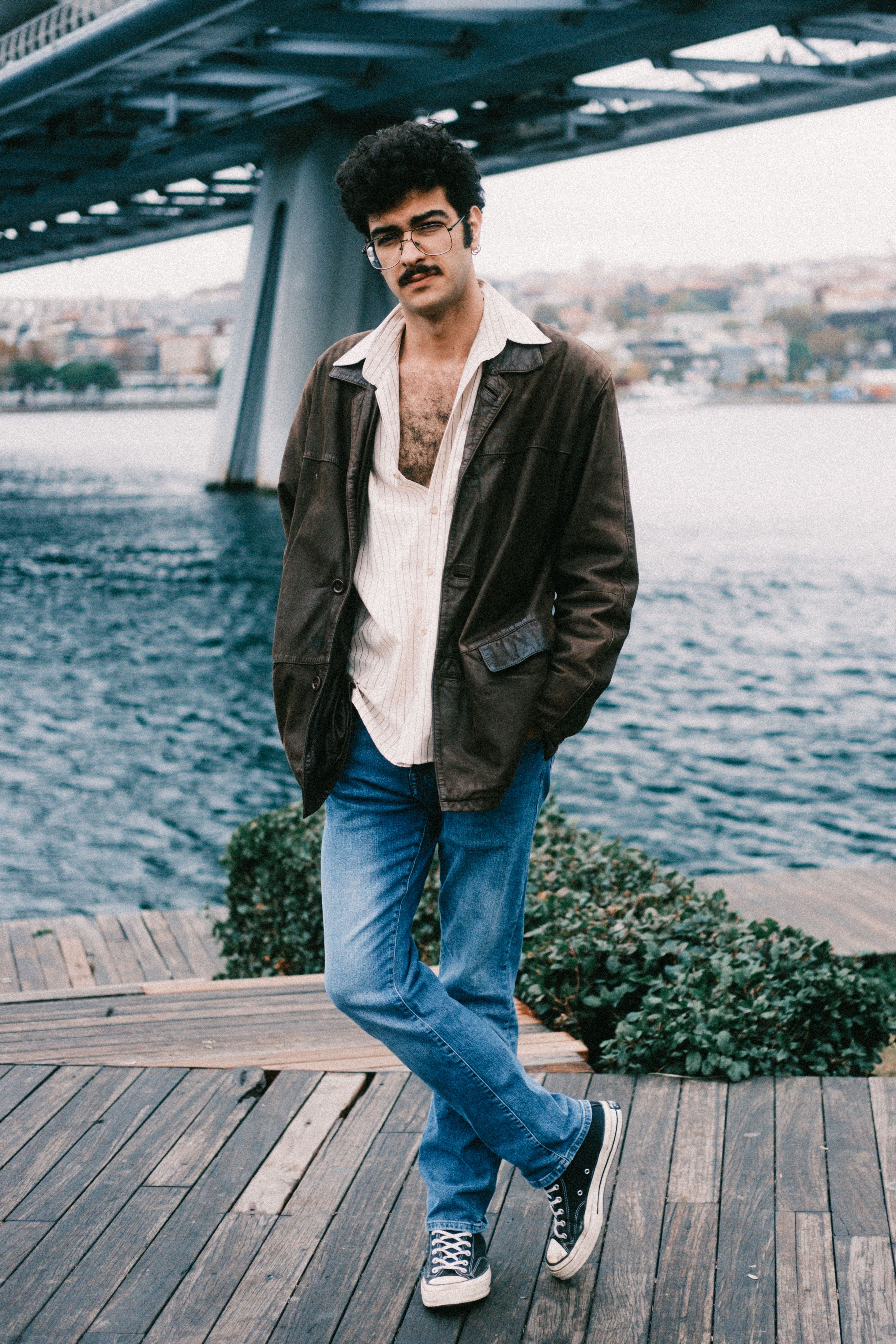 Young man in casual attire standing near a bridge over water, exuding a relaxed vibe.