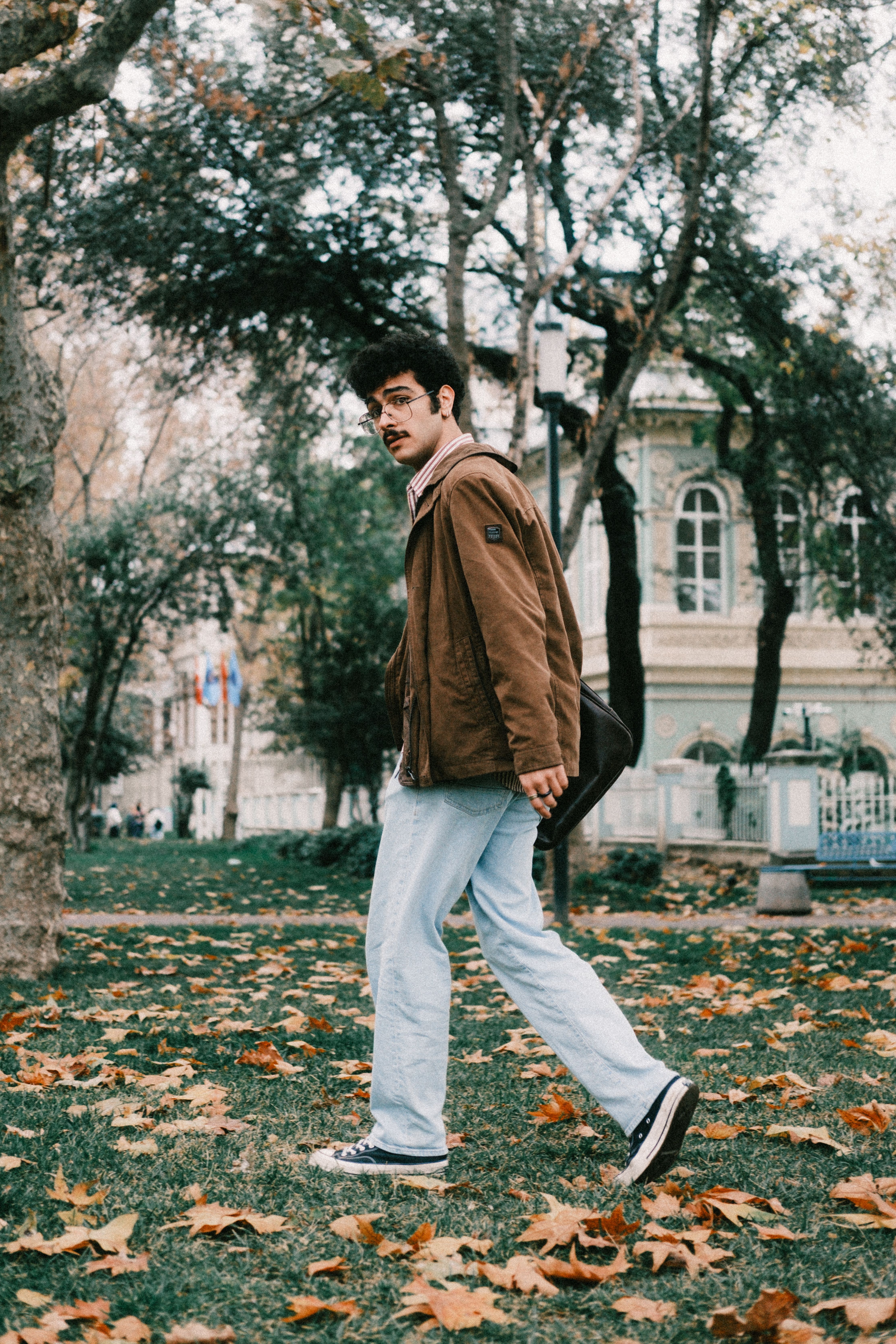 Casual adult male walking in a park with autumn leaves and vintage architecture.