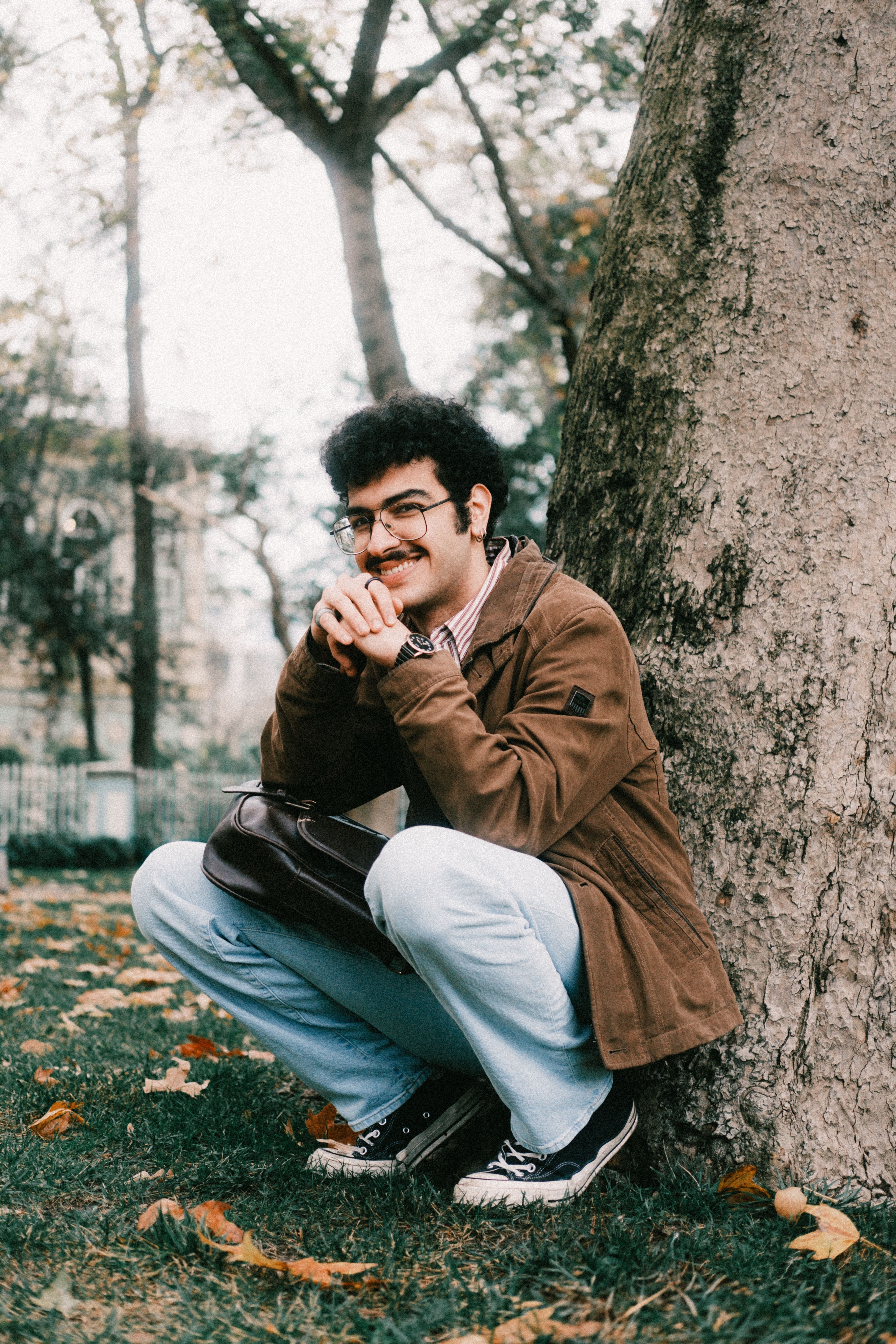 A young man in casual attire crouching by a tree in a serene park setting.