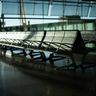 Rows of empty, shadowed seats in a quiet airport terminal with sunlight streaming through large windows.