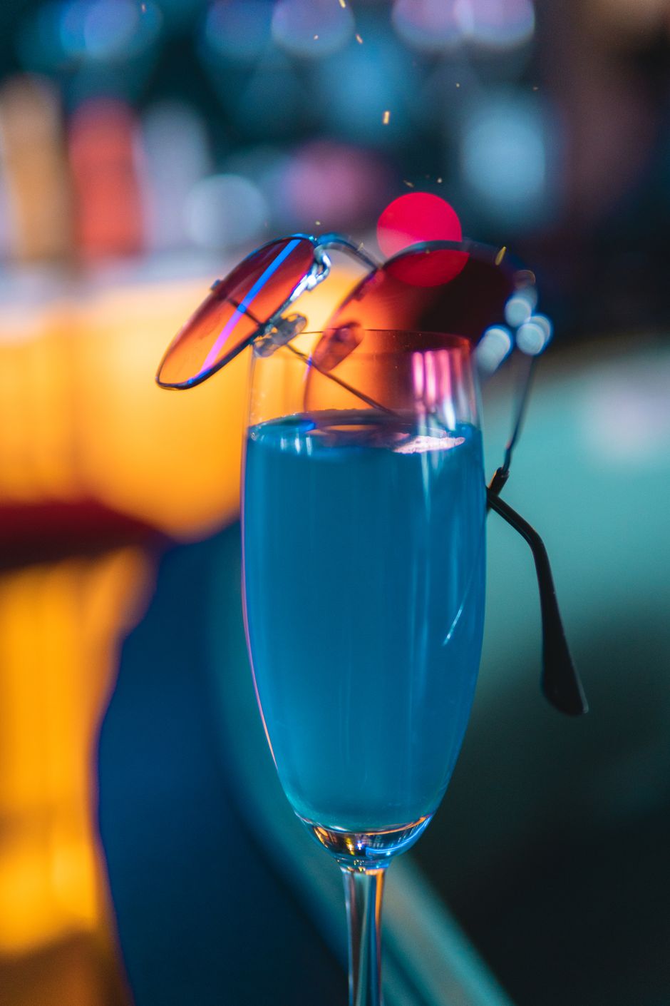 A stylish cocktail glass with a blue drink and sunglasses perched on top in a nightlife setting.