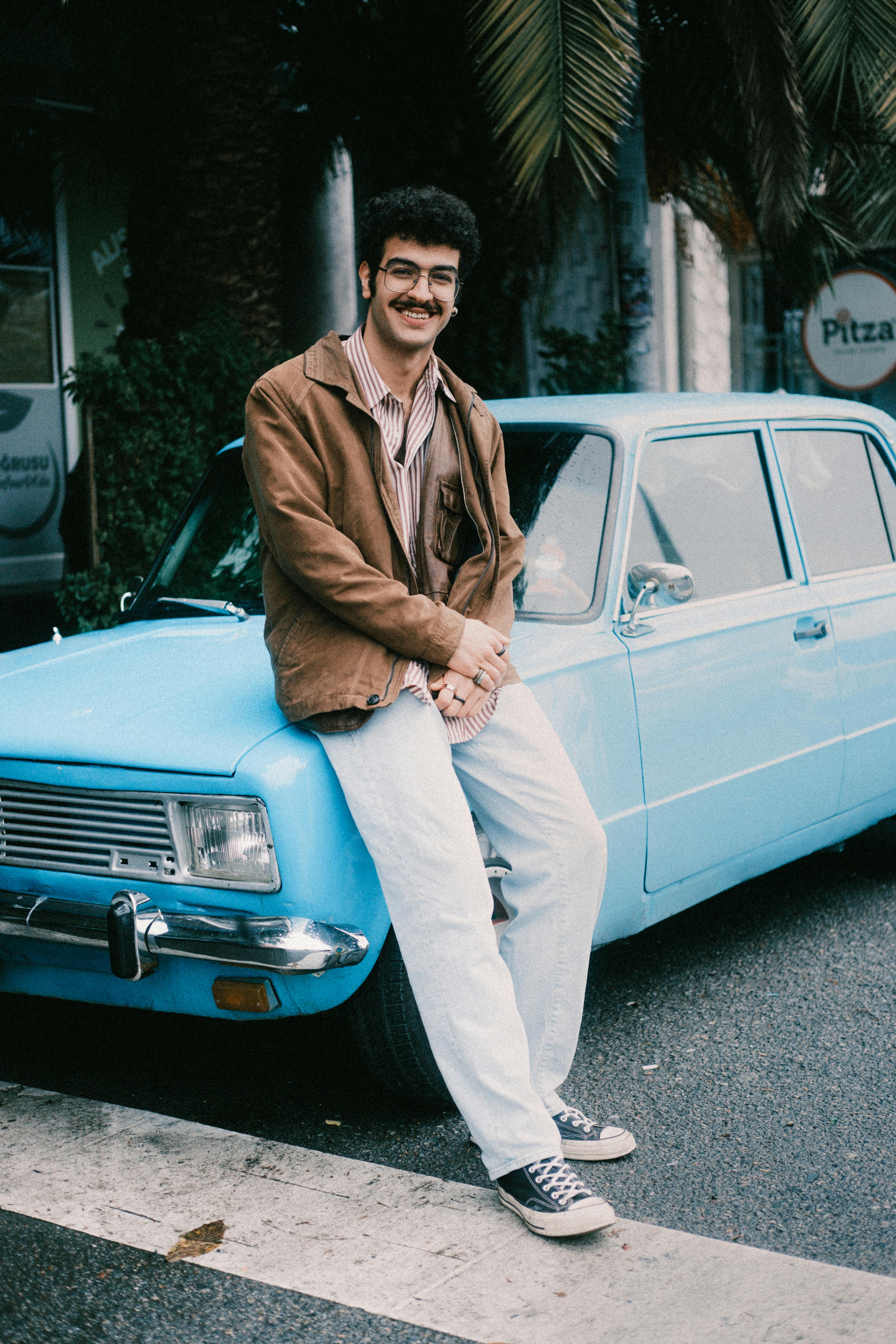 Man in casual attire leaning on a vintage blue car parked on urban street.