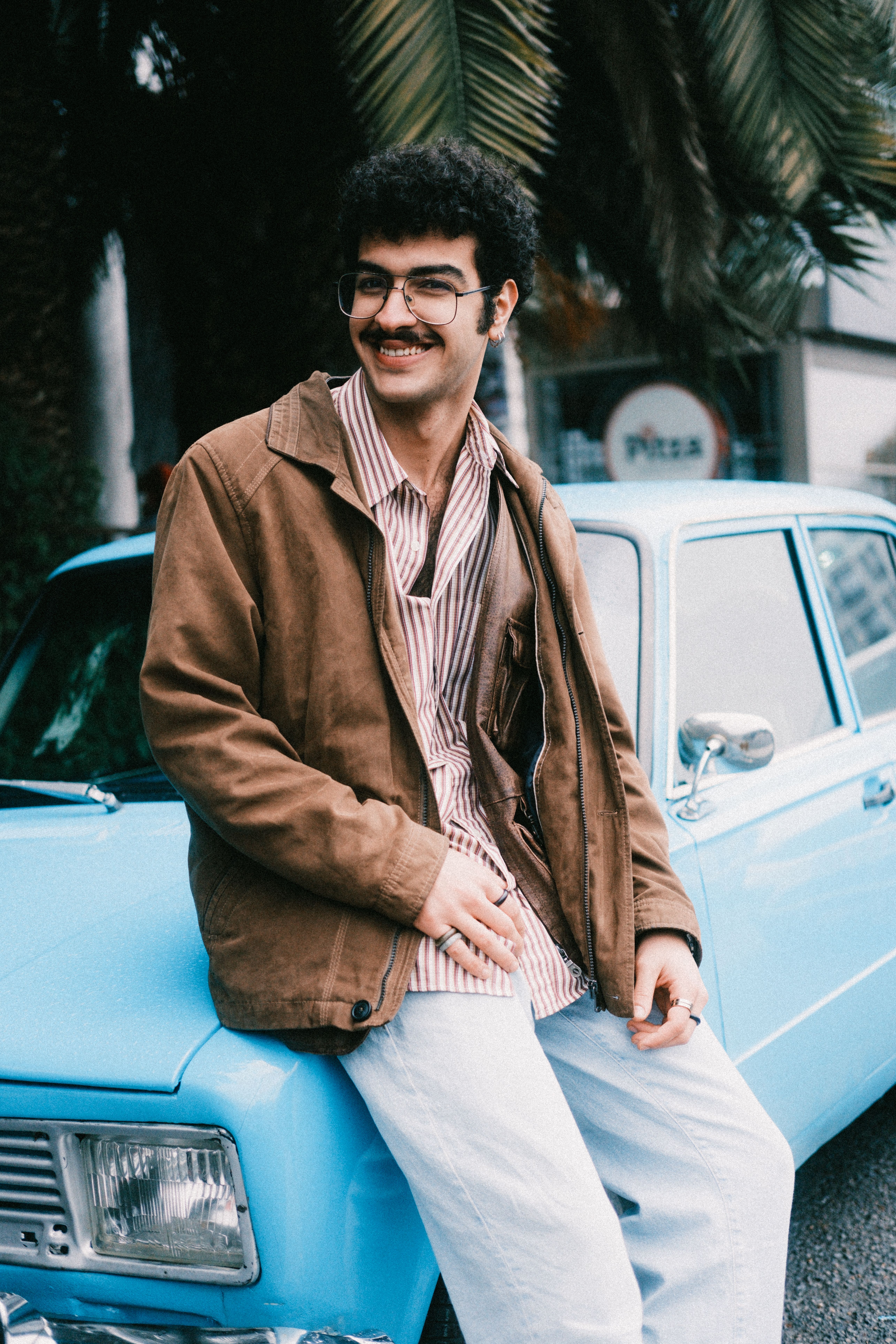 Smiling man in casual attire sitting on a blue vintage car surrounded by palm trees.