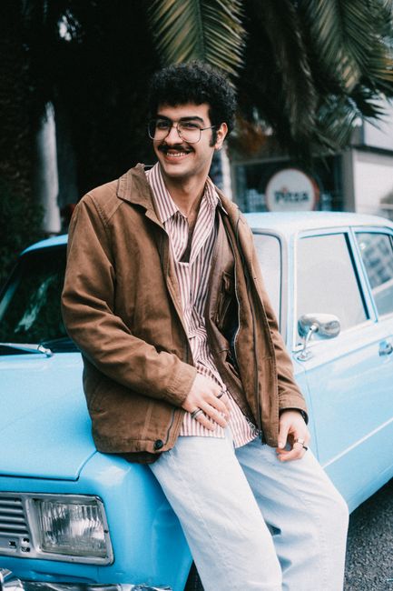 Smiling man in casual attire sitting on a blue vintage car surrounded by palm trees.