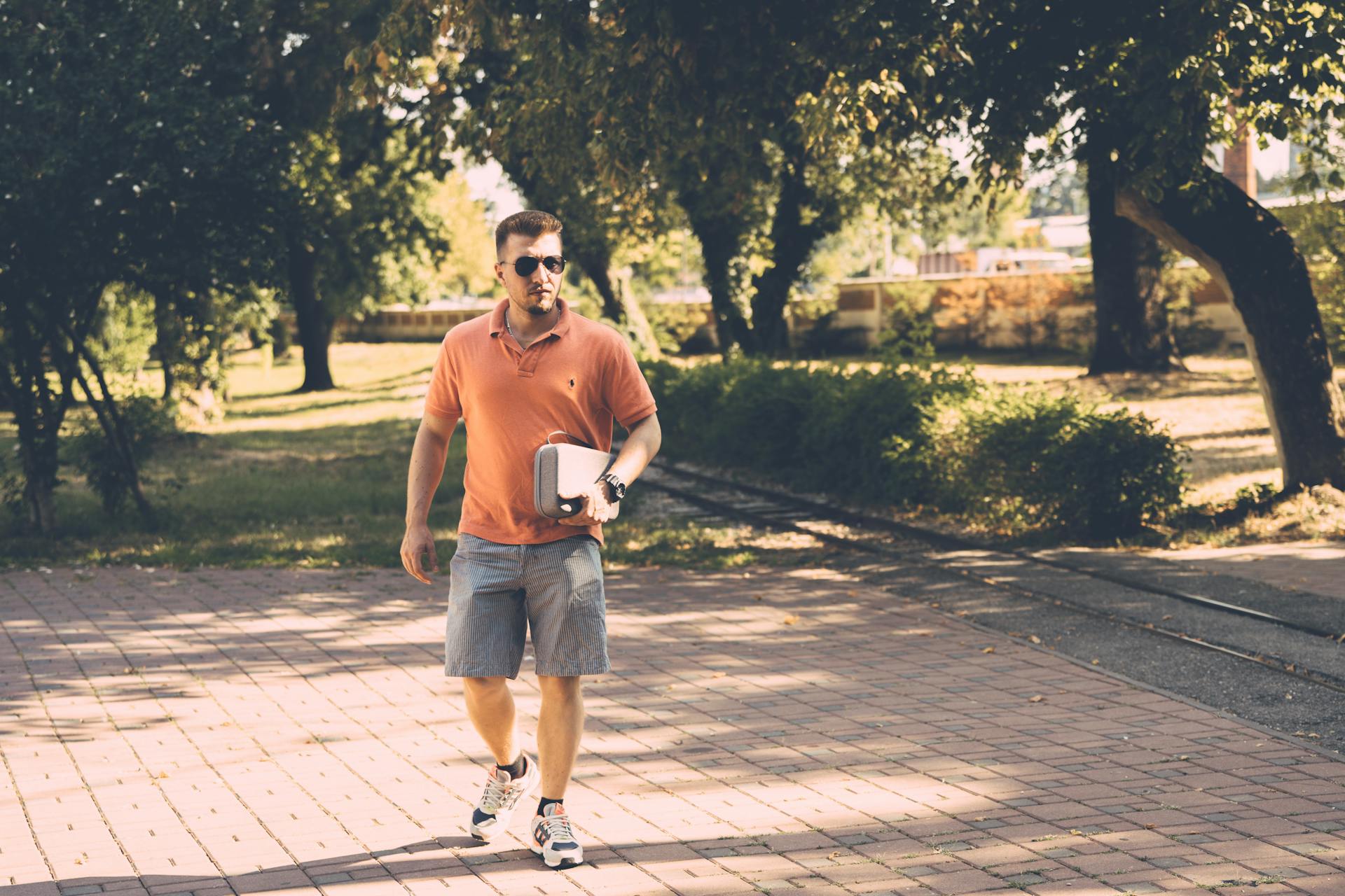 Casually dressed man with sunglasses carrying a portable cooler in a sunny park.