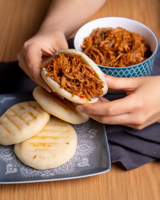 A split-open golden arepa on a wooden board, steam rising, filled with melted white cheese and shredded beef, with three more arepas cooling nearby.
