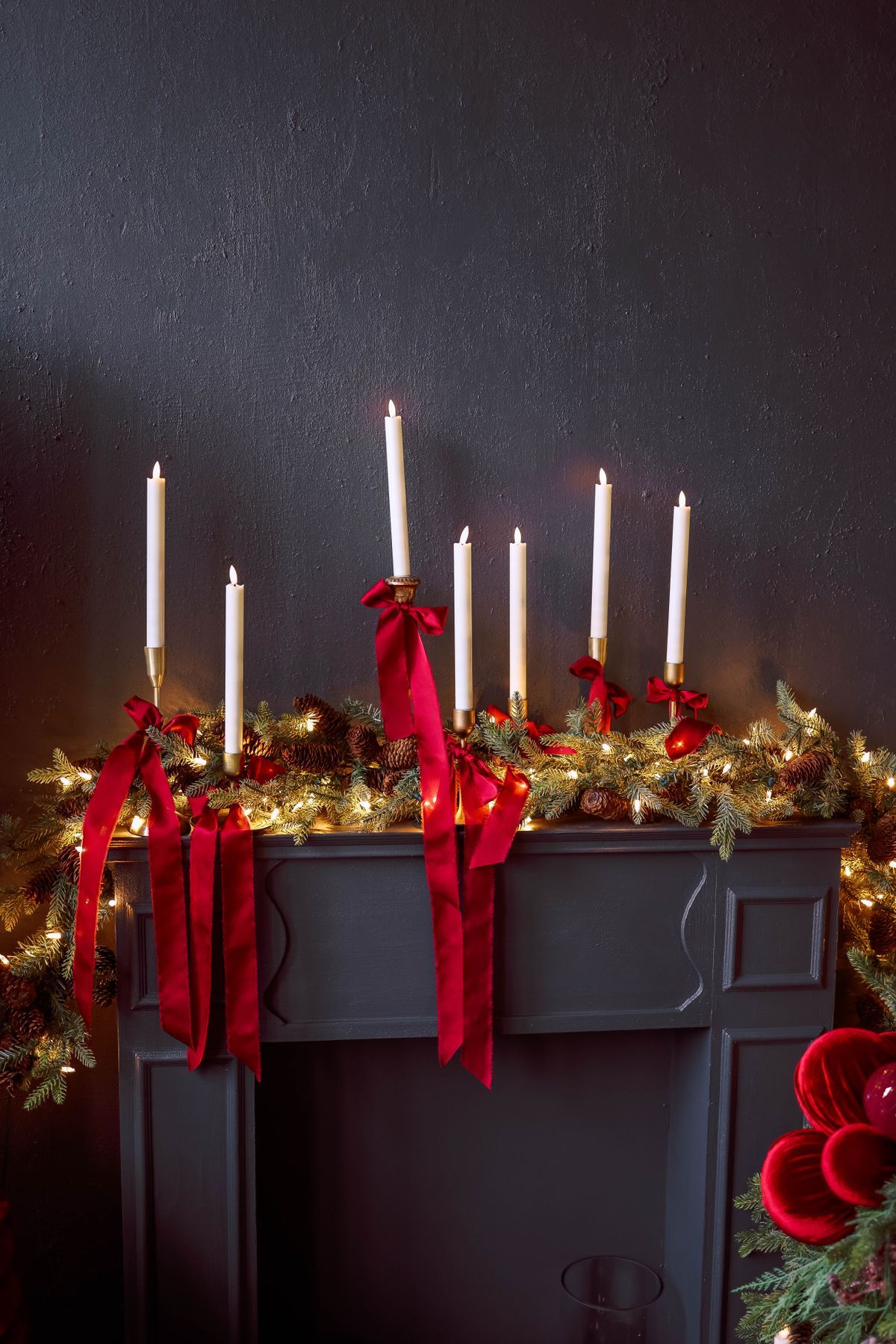 Row of tall white taper candles on a dark mantel with red velvet ribbon bows and a lit garland