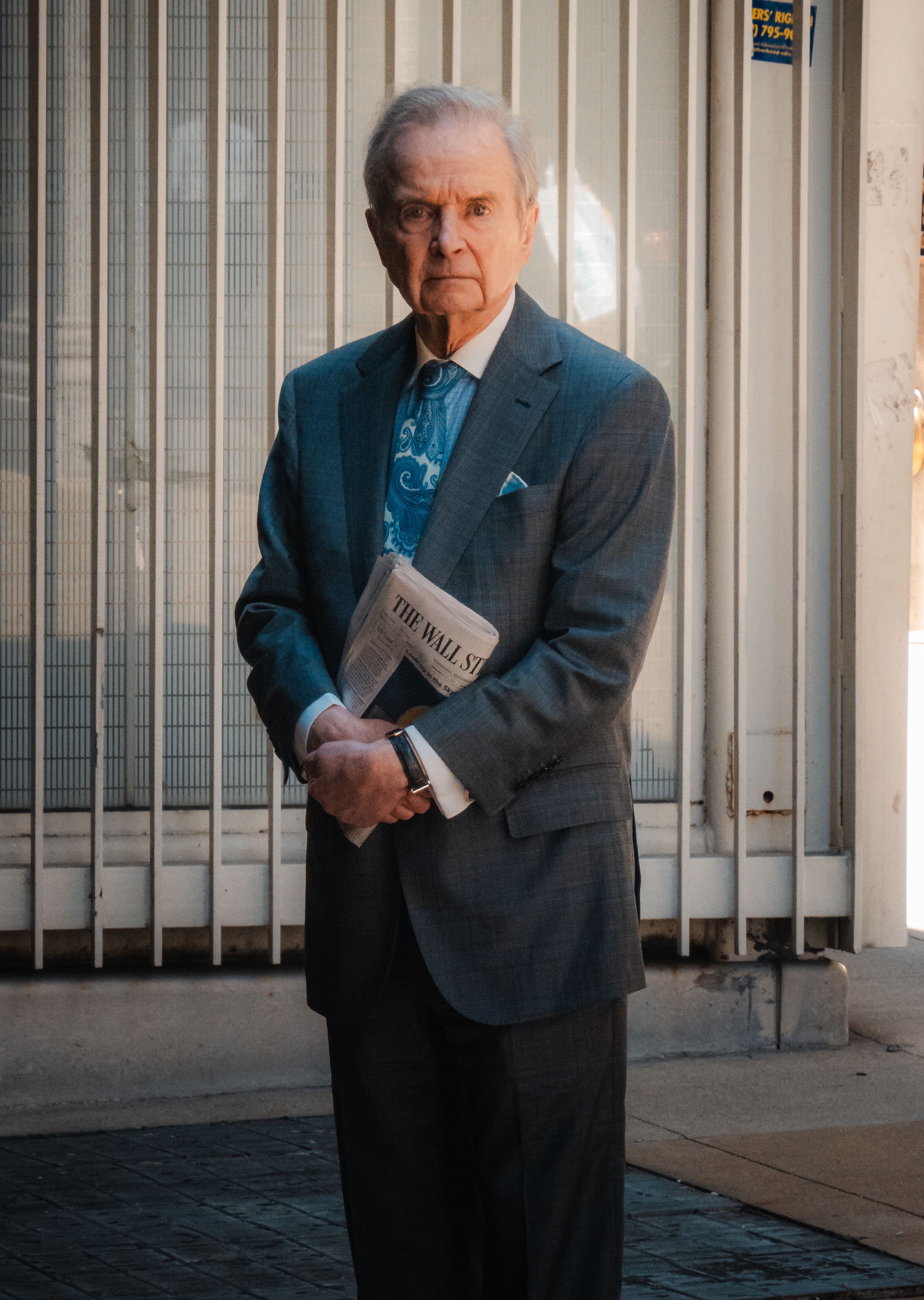 Elderly gentleman in a suit holding a newspaper outside a metal building.