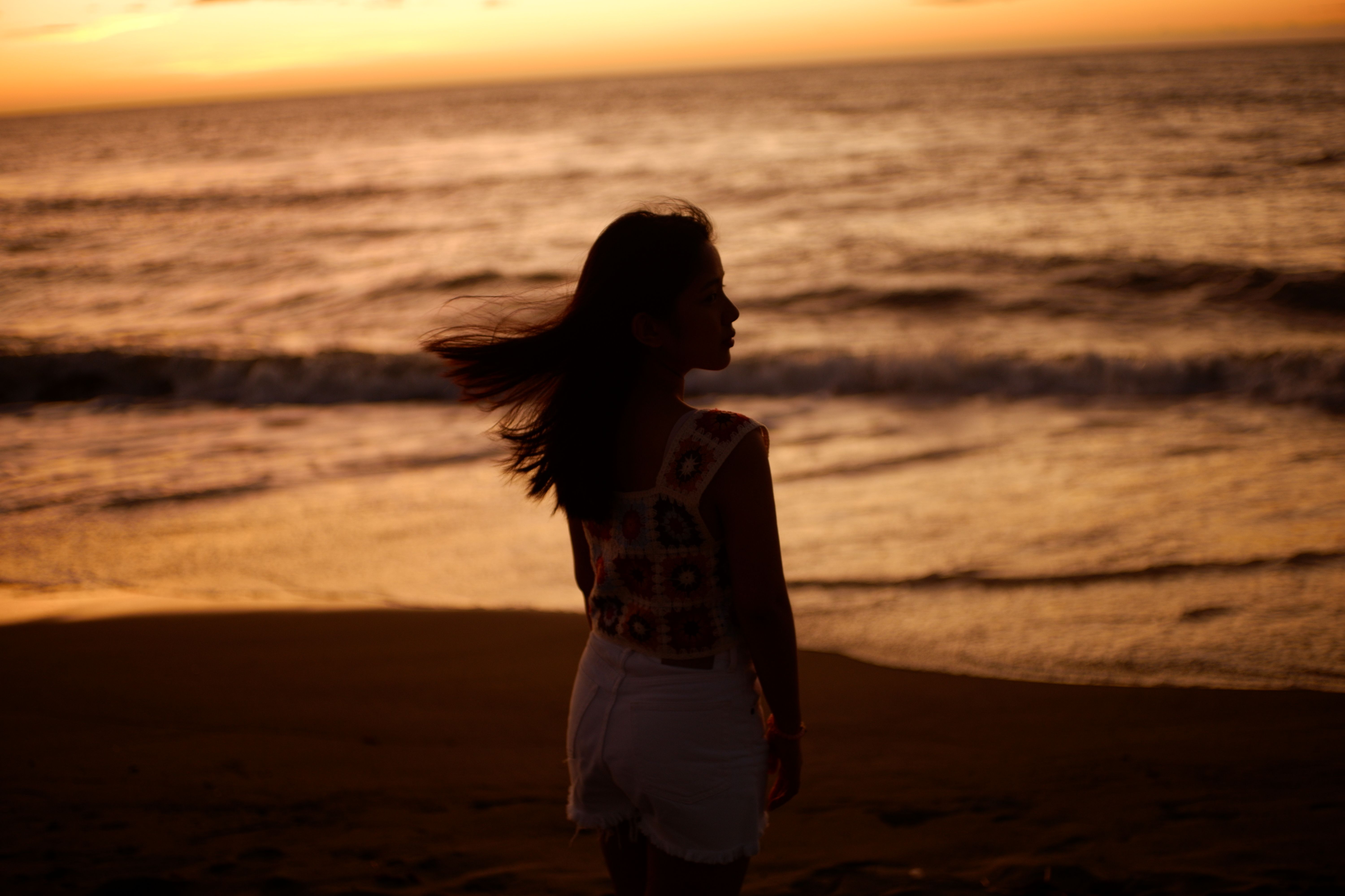 Gratis Silhouette di una donna su una spiaggia ventosa al tramonto, che cattura serenità e bellezza. Foto a disposizione