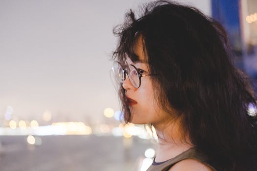 一位亚洲女性戴着眼镜、留着长发，她的侧面轮廓映衬在散景的城市灯光下