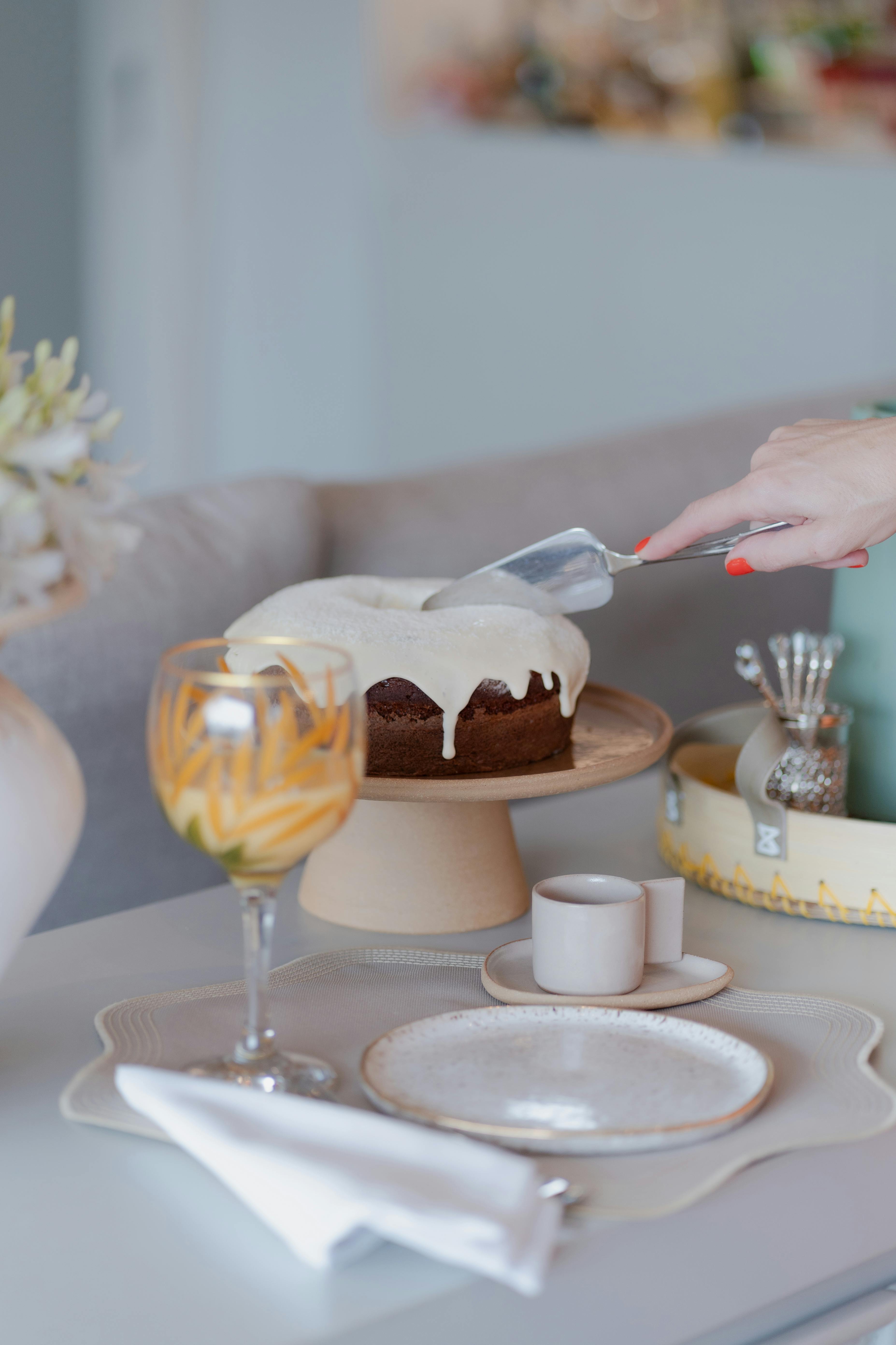 A hand slices a frosted cake on an elegant dining table set with plates and glasses.
