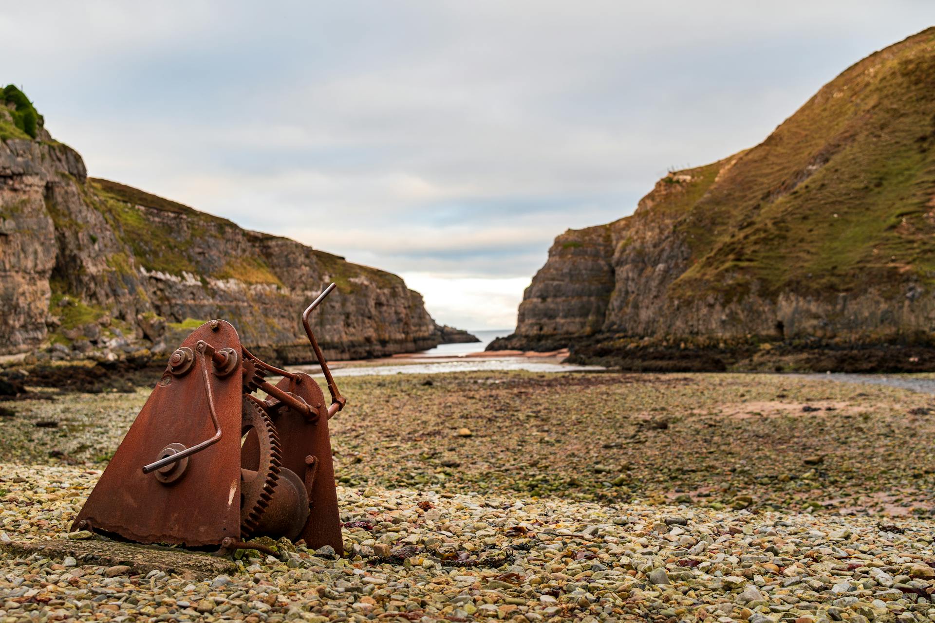 Rusty mechanical gear on rocky beach with scenic seaside cliffs, ideal for industrial and nature themes.