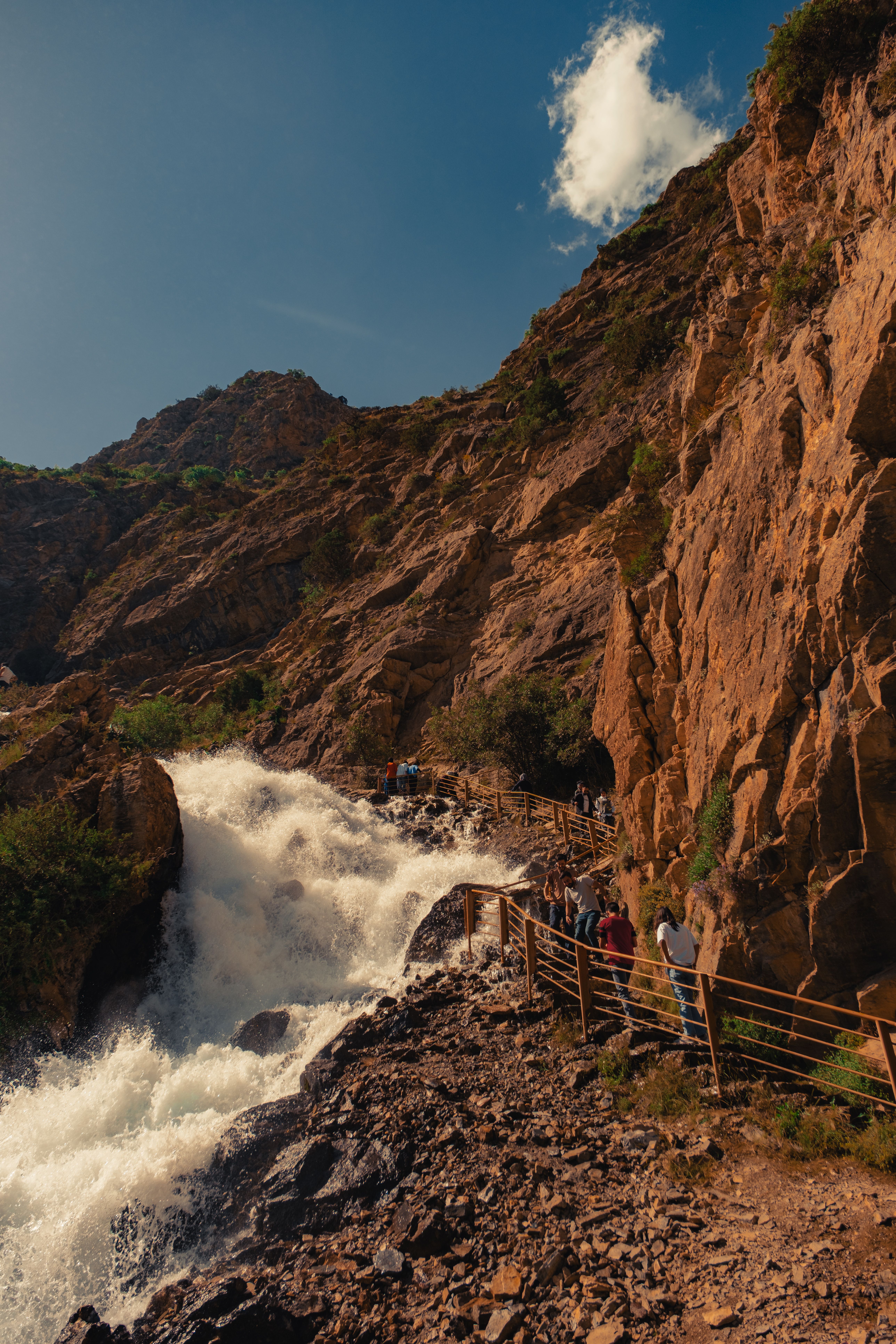 Explore nature with hikers on a scenic trail beside a roaring waterfall surrounded by rugged cliffs.