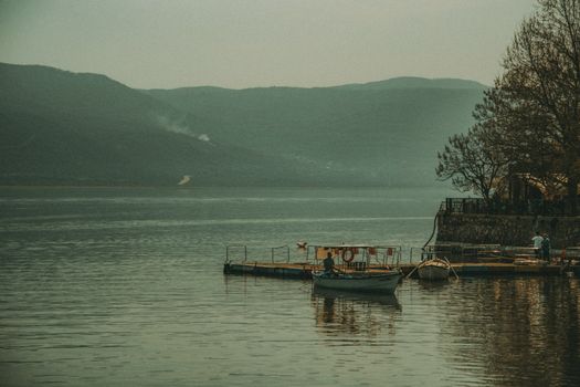 A peaceful lake view with a wooden pier, boats, and distant mountains under a muted sky.