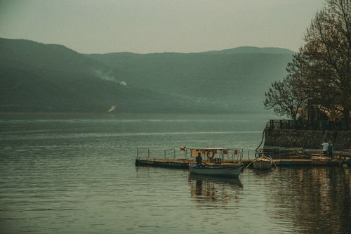 Free A peaceful lake view with a wooden pier, boats, and distant mountains under a muted sky. Stock Photo