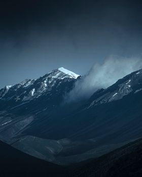 Majestic mountain range with snow-covered peak and dramatic clouds under a moody sky.