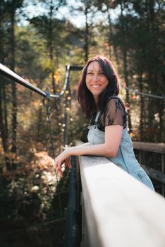 A woman in a casual outfit smiles on a wooden bridge under a sunlit forest canopy.