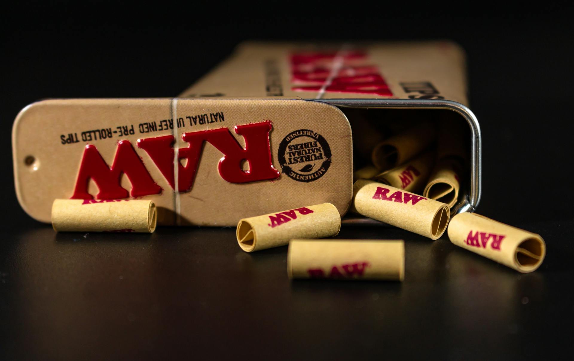 RAW pre-rolled tips spilling from a metal tin on a dark surface, close-up view.