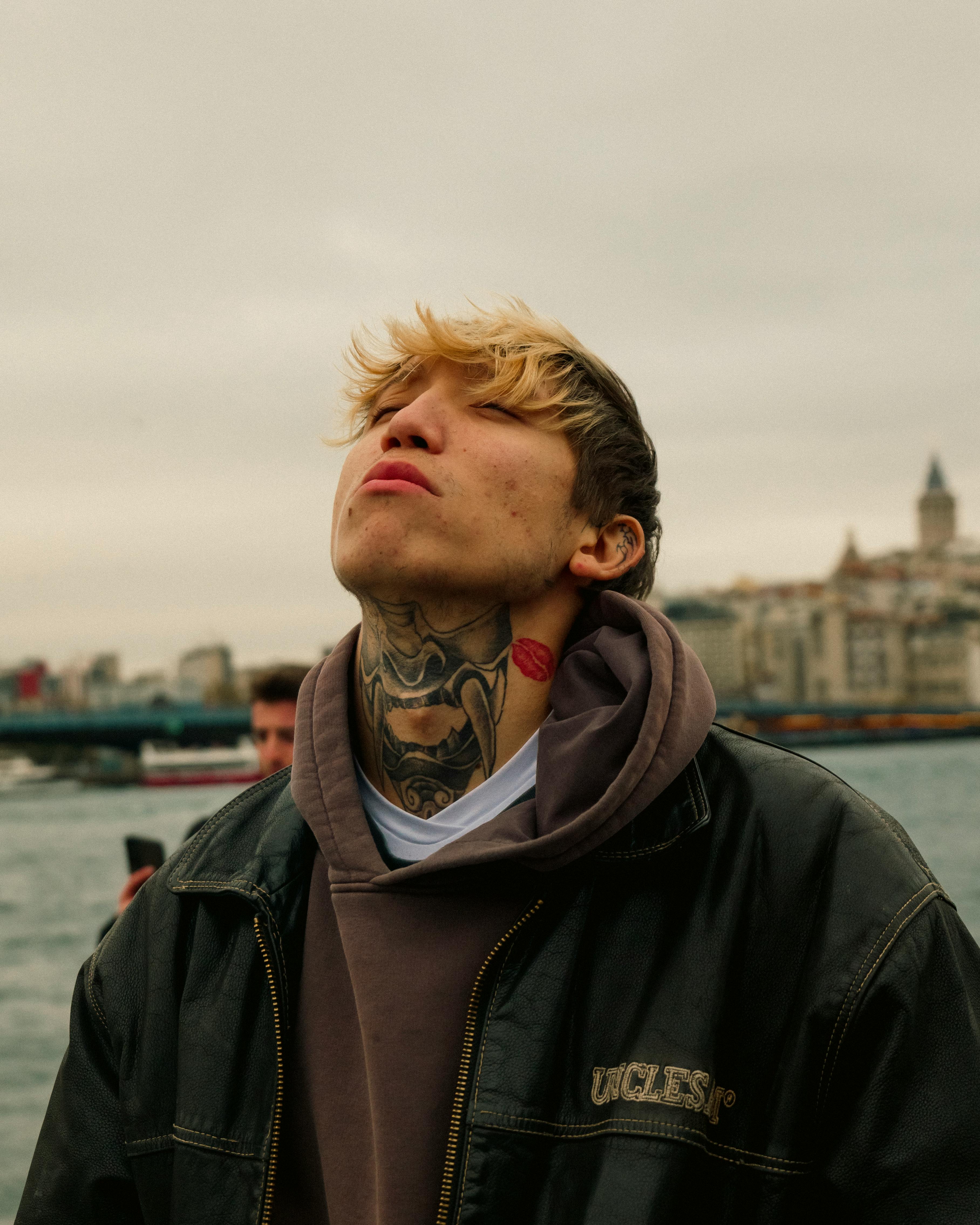 A tattooed man in a hoodie enjoys the breeze near the waterfront with urban scenery behind.