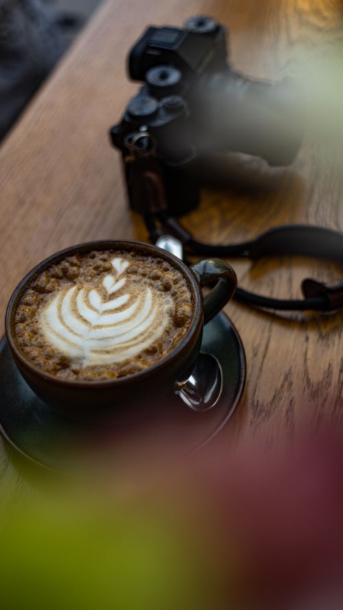 Free A latte with elegant latte art sits beside a camera on a wooden table, embodying creativity in Ankara. Stock Photo