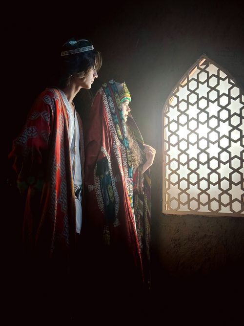 Free A couple in traditional clothing gazes through a lattice window, illuminated by warm light. Stock Photo