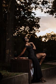 Stylish woman in a black dress posing elegantly outdoors at sunset.