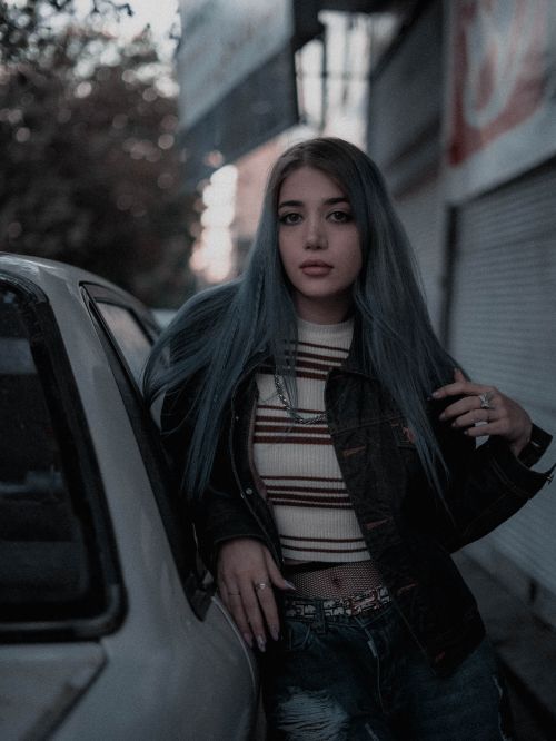 Free A fashionable young woman with blue hair poses confidently beside a car in an urban environment. Stock Photo