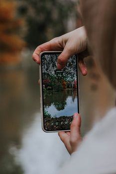 A person photographs autumn scenery with a smartphone by a lake.