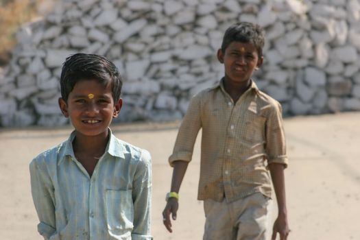 Two boys outdoors, one smiling, against a pile of white stones on a sunny day.