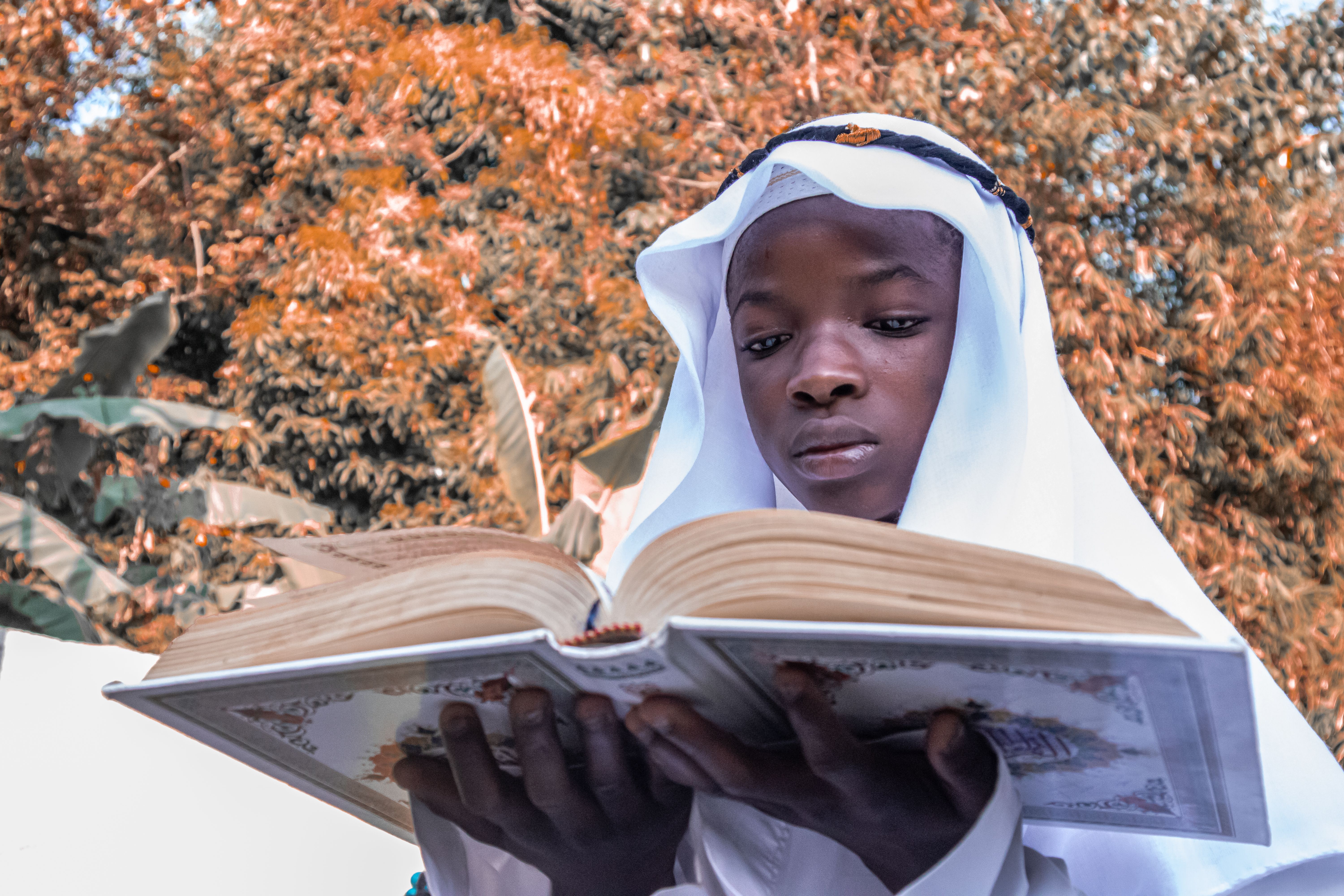 Boy in traditional Islamic attire reading Quran, with autumn foliage in the background.