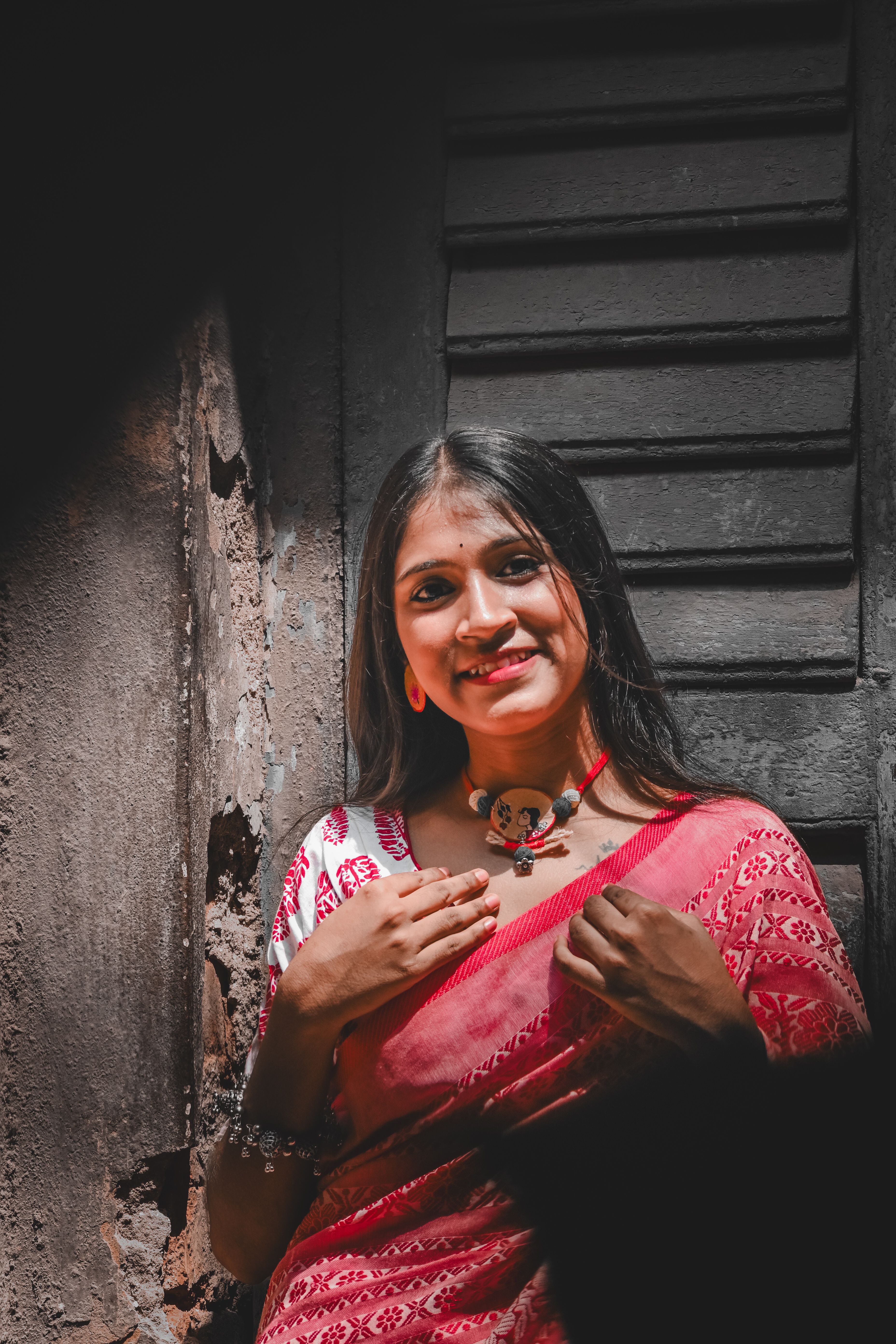 Young woman in a vibrant red saree standing by a rustic wooden window, smiling warmly.