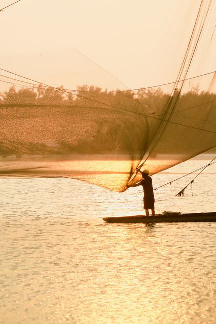 A silhouetted fisherman skillfully casts a net over a tranquil water body during sunset, capturing the serene beauty of traditional fishing.