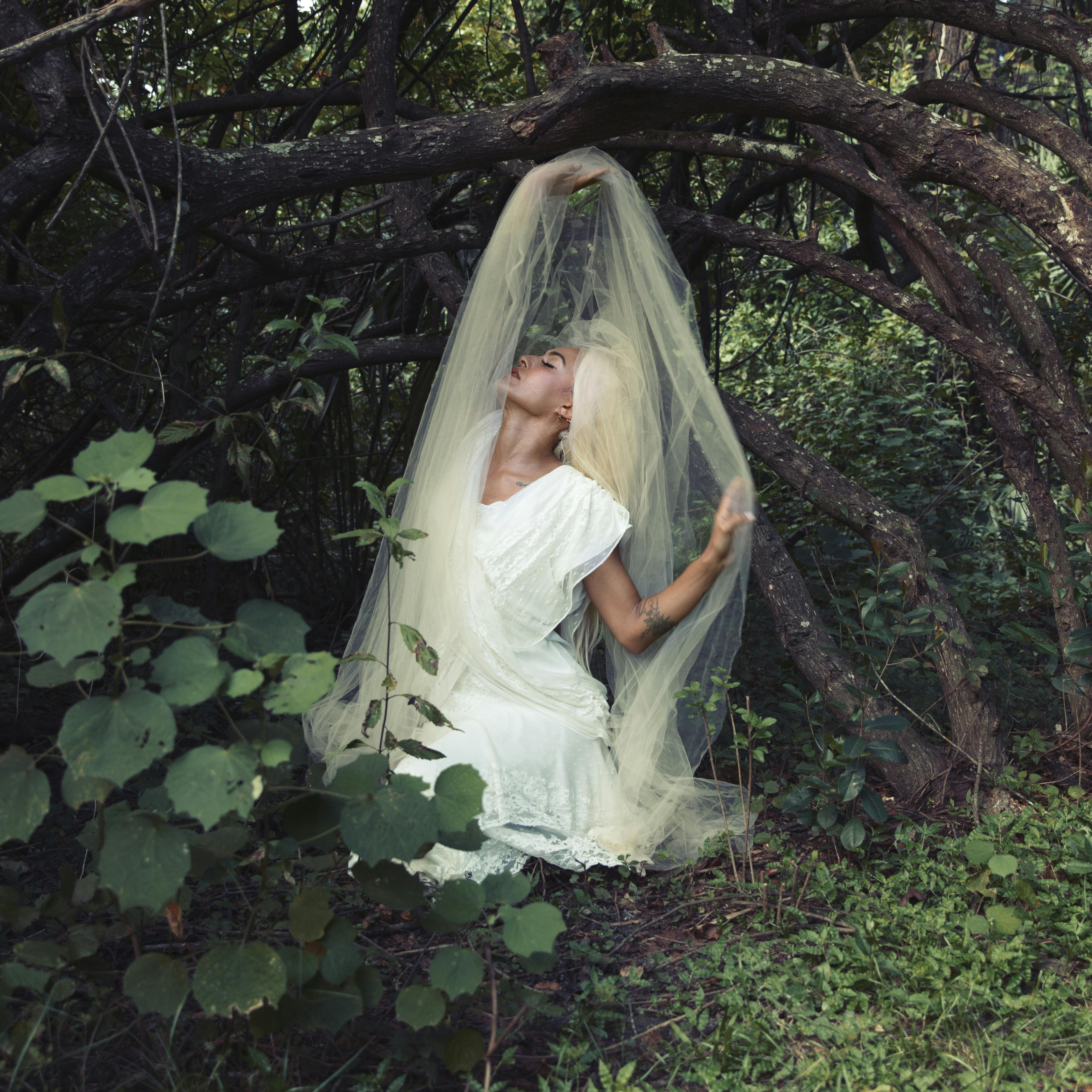 A woman in a white dress and veil poses gracefully in a mystical forest setting.