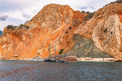 Free Vibrant view of boats docked by red cliffs with tourists on the beach under a bright sky. Stock Photo