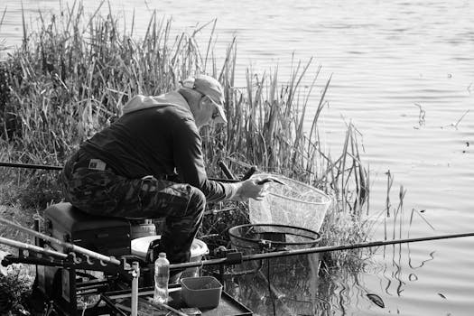 A black and white photo of an elderly man fishing by the riverbank, enjoying a peaceful day.