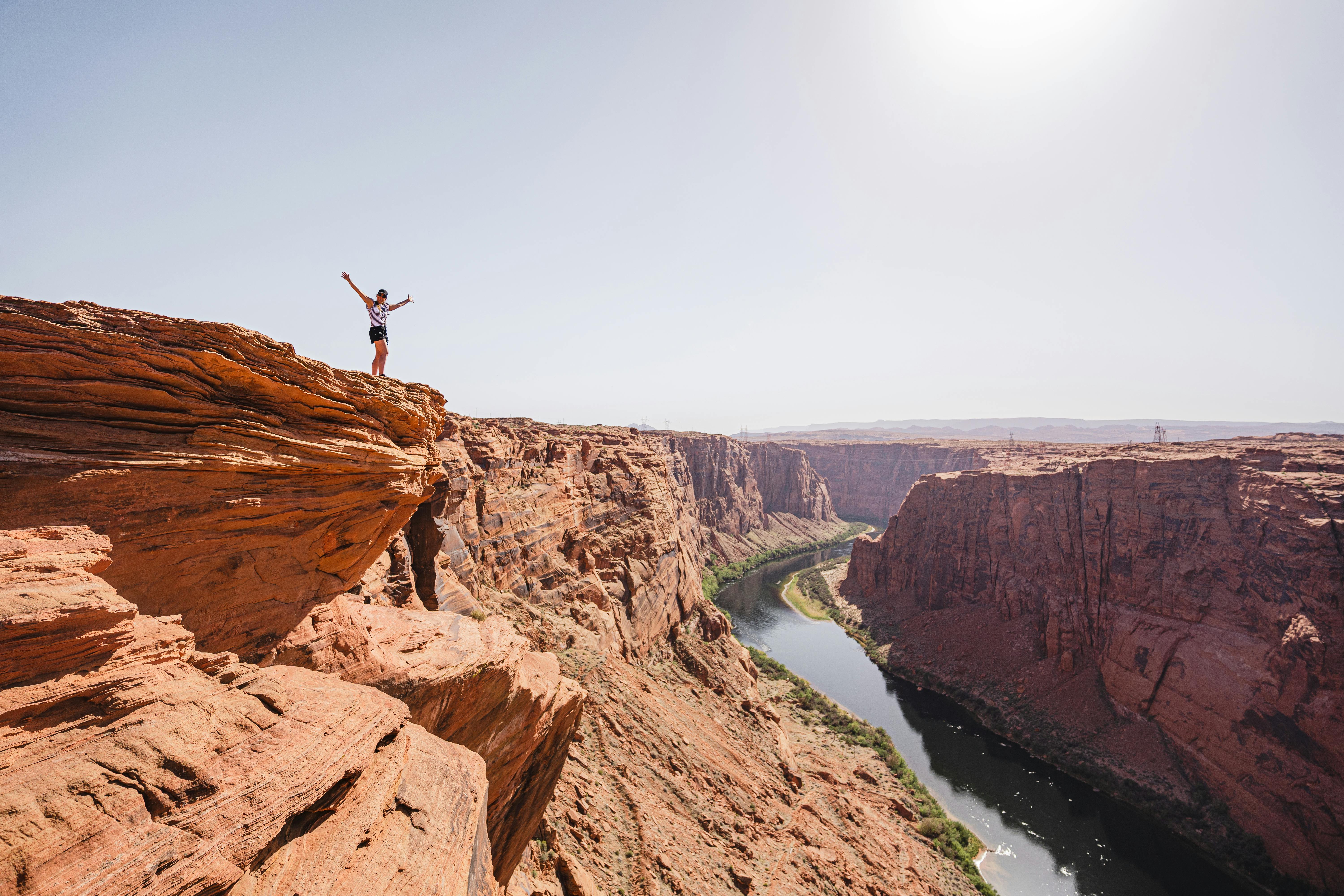 A hiker stands triumphantly on the edge of Horseshoe Bend in Page, Arizona under a bright sun.