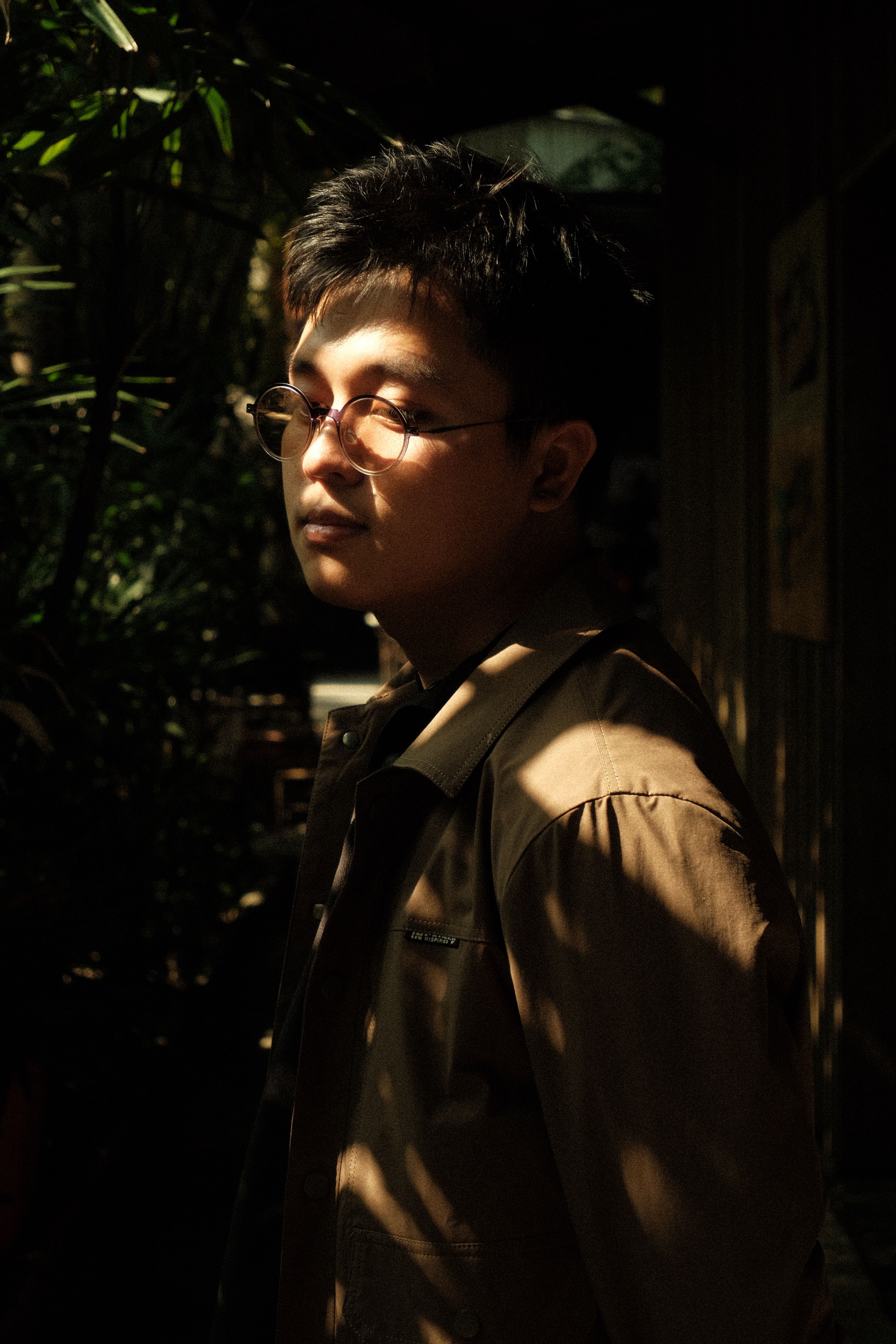 A young man in thoughtful pose with glasses, enveloped by dappled sunlight in a lush setting.