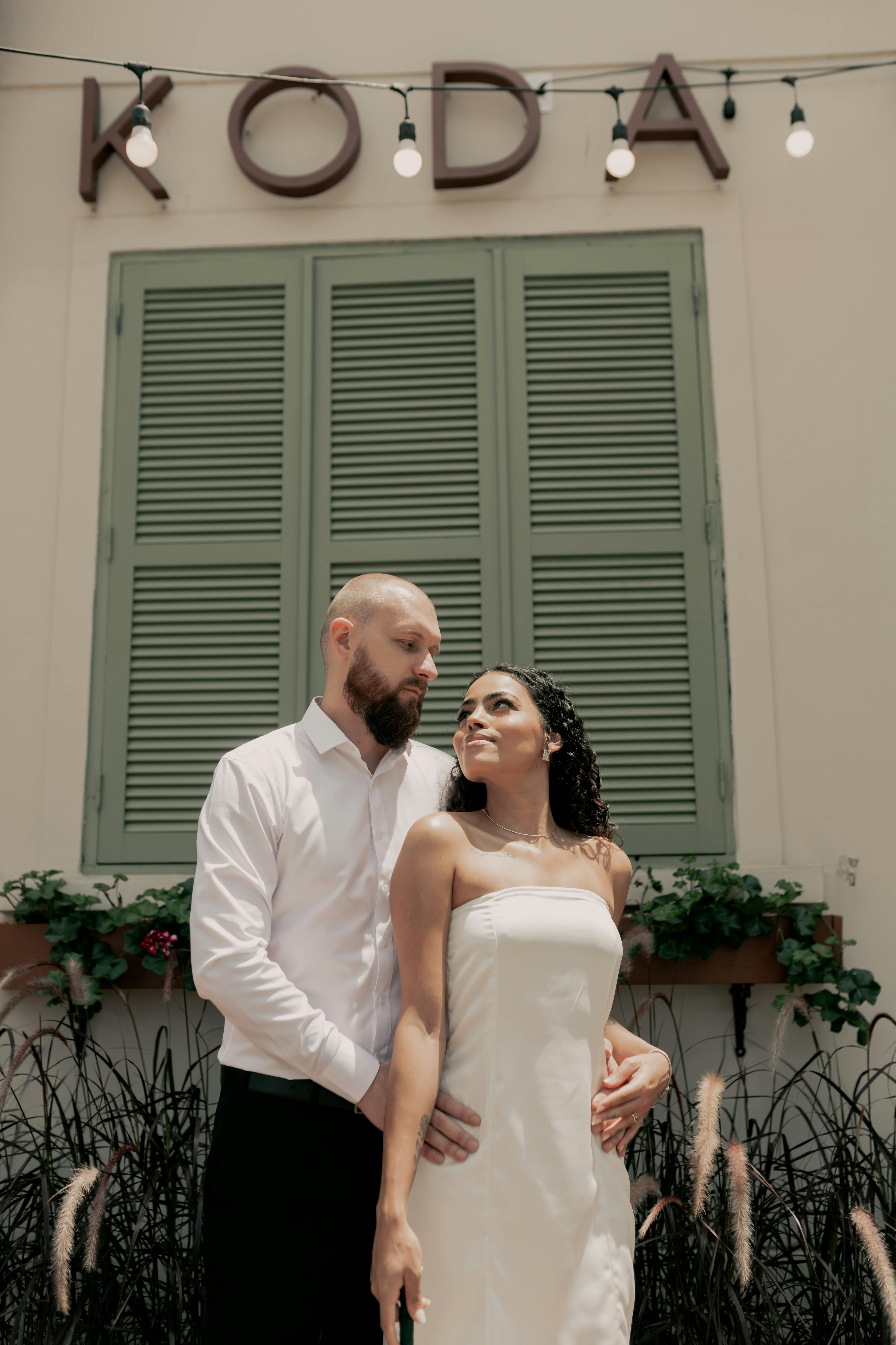 A couple embraces in front of a café with greenery and string lights.