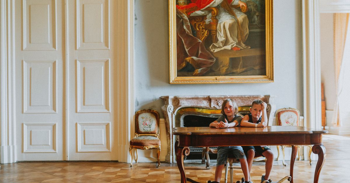 Two children sit at an antique table beneath a grand historical painting in an elegant room.