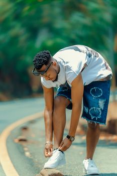 Casual young man tying shoelace on a scenic outdoor road, wearing denim shorts and sunglasses.