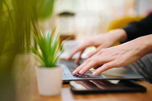 Hands typing on a laptop in an indoor setting with a green plant. Perfect for work from home concepts.